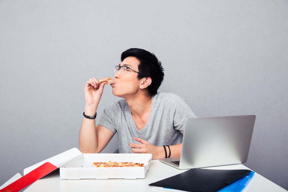 Handsome asian man sitting at the table and eating pizza over gray background